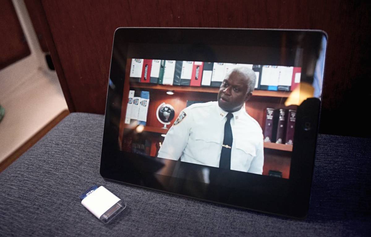 An Ipad2 is laying on its side, held up by its magnetic cover(which is folded up into a shape resembling an extruded triangle, forming a stand). On screen, an episode of Brooklyn Nine Nine is playing, with Captain Holt in his office.
A camera connector with an SD card inserted into its slot is lying nearby.