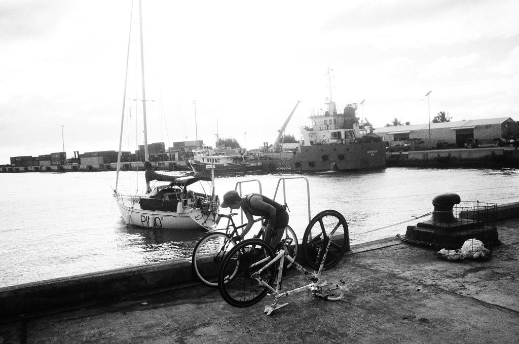 Rek building the bikes while in Avatiu Harbor in Rarotonga in the Cook Islands A black and white photograph of someone putting two bikes together on a concrete pier overlooking a harbour with a sailboat med-moored in the background
