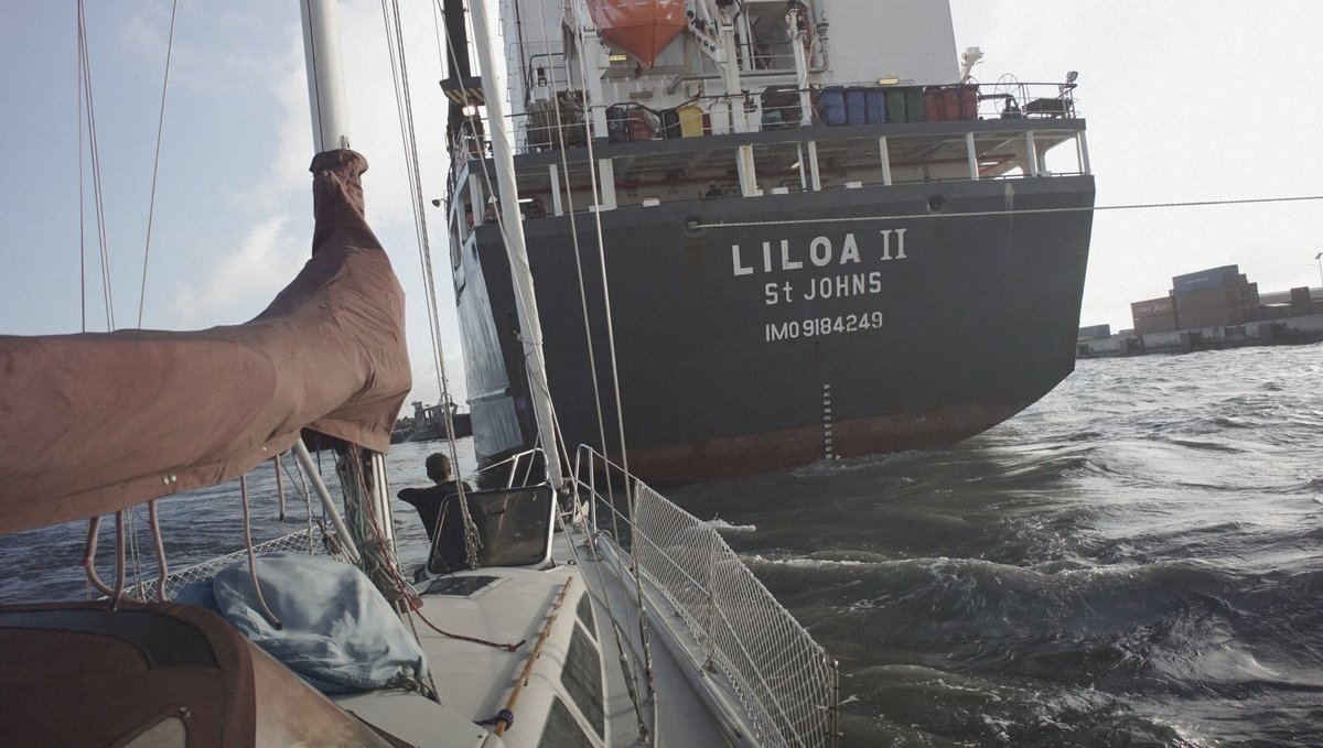 A cargo ship backing up too close to Pino while in Avatiu Harbor Rek sitting on the bow of a sailboat while moored into a port with the stern of a large cargo ship just a few short feet away as it is trying to dock, its powerful propellers raising wakes