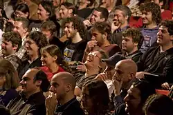 About 20 people in a crowd, reacting to an unseen speaker. Some are laughing, some look very serious.