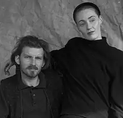 Man and woman with dark hair, both in dark clothing against plain background