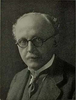 Black-and-white portrait of a bespectacled, balding man with a moustache