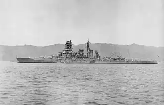 Side view of an anchored dark-gray warship with four gun turrets, two funnels, one mast and a prominent forward superstructure.