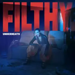 A photograph of Shah staring ahead while seated on a lounge in a room illuminated with a blue glow, wearing a tight black top, baggy pants and high heels