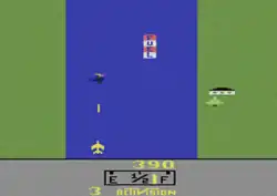 A rectangular video game screenshot that is a digital representation of a jet fighter over a river and two grass-filled shores. The jet fighter is shown firing a missile at a helicopter while a tree and a dwelling are displayed on the grassy portion. A tank labeled "Fuel" is shown resting on the water.