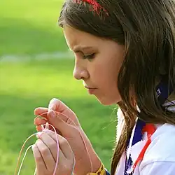 A girl with long brown hair concentrates on threading pink string though white string, on a sports field