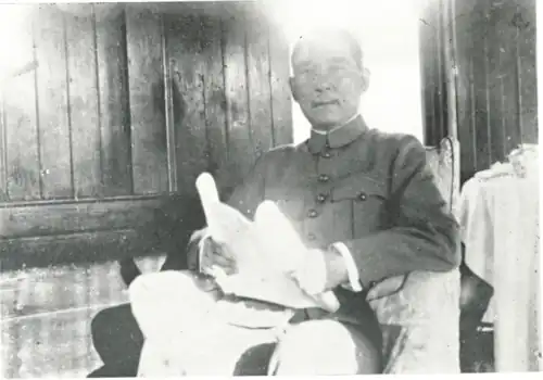 A black and white photograph of a seated man wearing a military or pseudo-military jacket