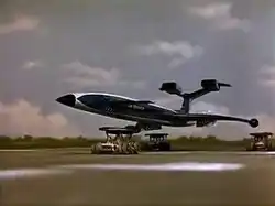 A blue, futuristic airliner rests on top of three orange support vehicles (one underneath the fuselage and two underneath the wings) as it speeds out of control down an airport runway, the sky bright with clouds.