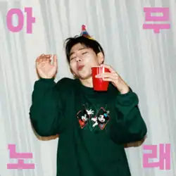 A man wearing a green shirt and a birthday hat standing with his eyes closed against a gray-tinged background. Both his arms are raised to the height of his shoulders and he holds a red coloured plastic cup in his left hand.
